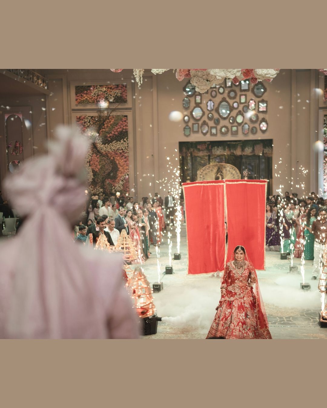A bride in red lehenga walks down a grand aisle with sparklers and floral decor at an Indian wedding ceremony.