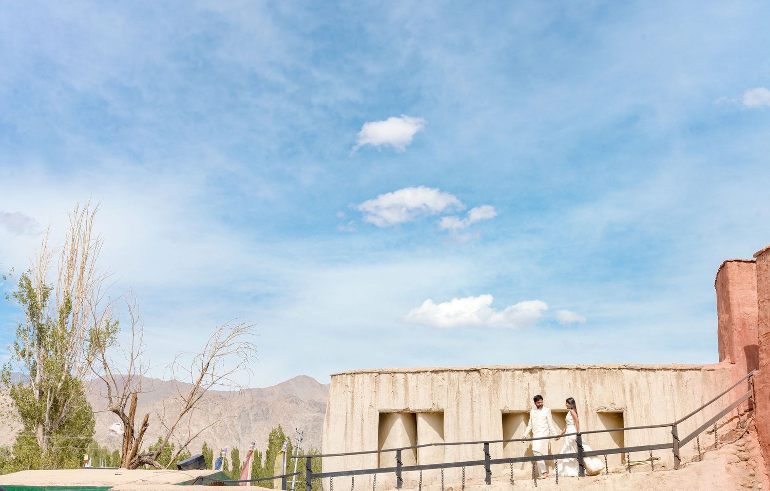 Couple in white attire on a ramp at a desert adobe building under a blue sky with mountains behind.