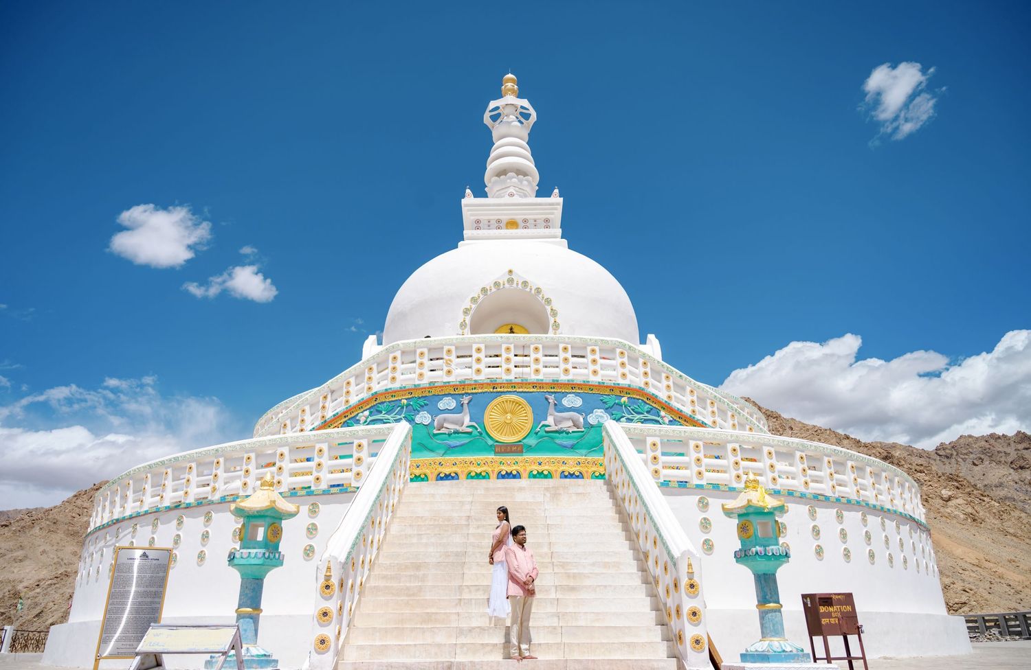 Couple standing on steps of Shanti Stupa, a white Buddhist stupa in Leh, Ladakh, India under a bright blue sky.