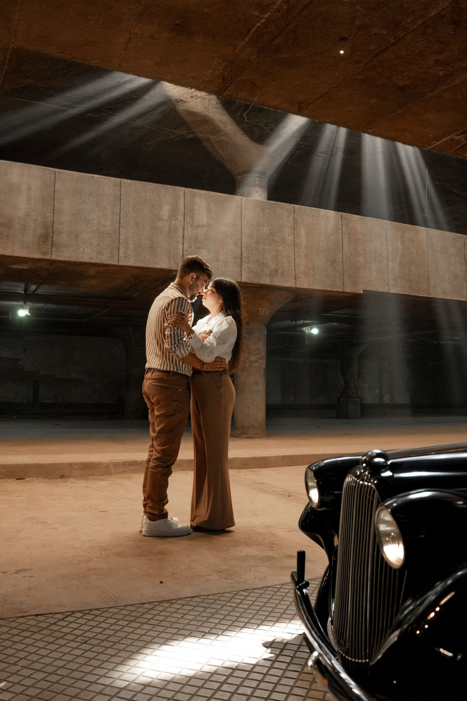 Couple embracing romantically in dramatic underground parking garage with vintage black car and light rays.