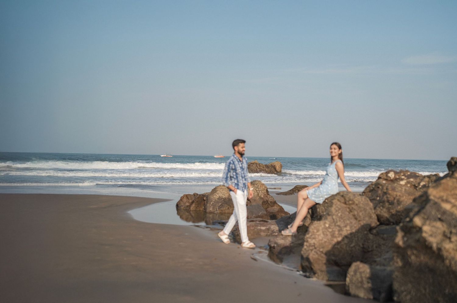 A couple walks along a rocky beach shore with ocean waves in the background on a clear sunny day.