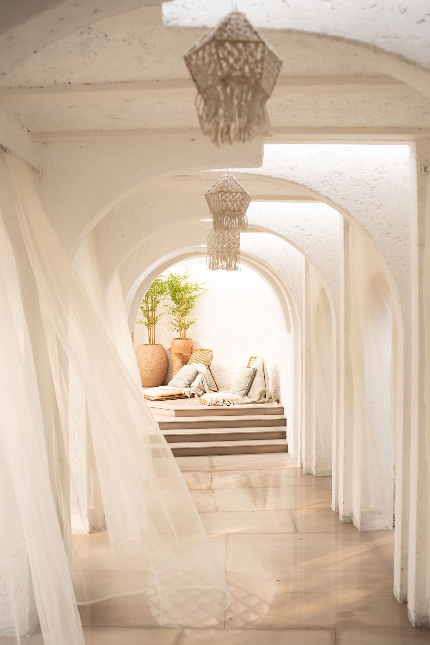 Elegant white arched corridor with sheer curtains, macrame chandeliers, terracotta pots, and a cozy seating area in warm natural light.