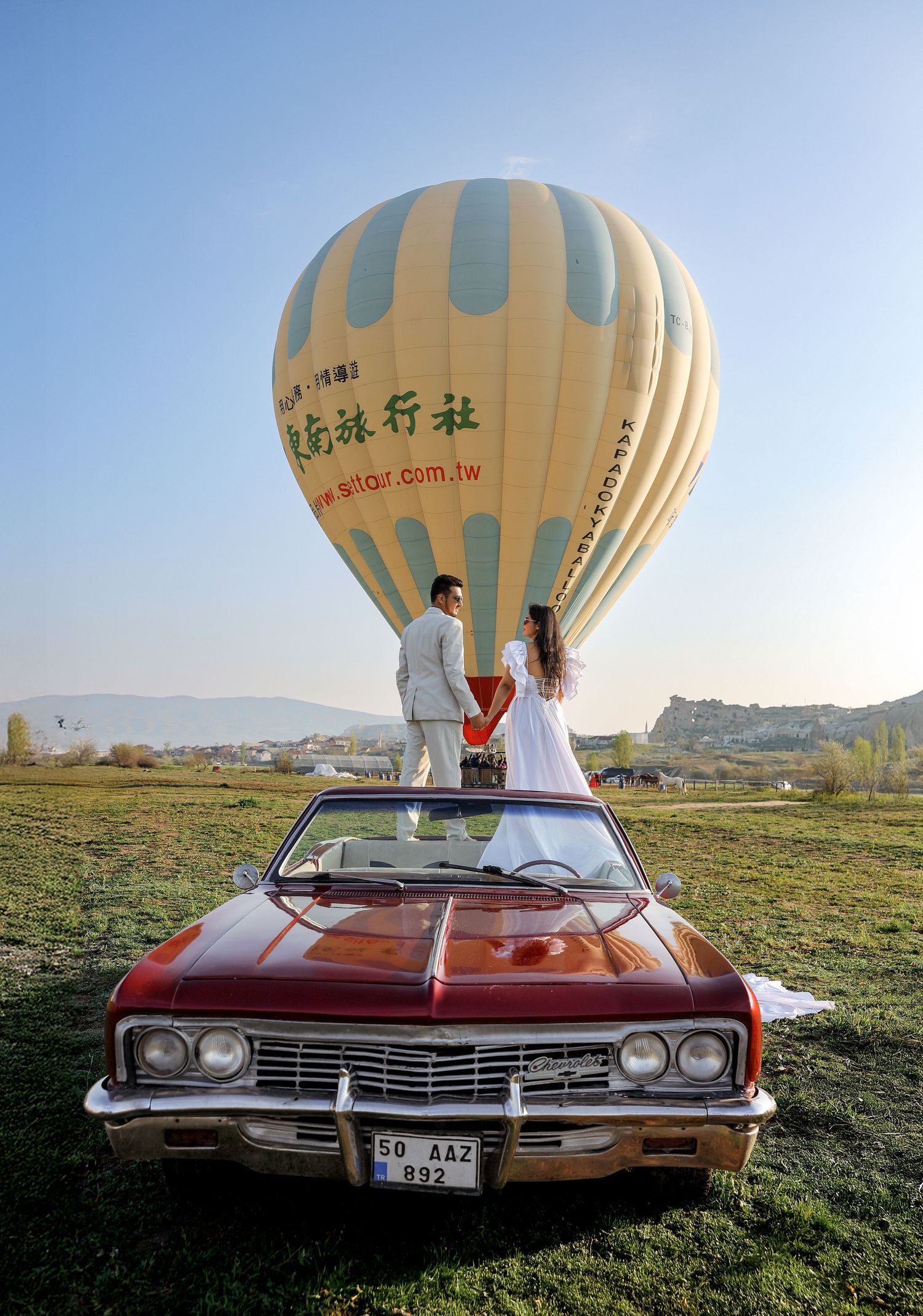 Vintage red car parked beneath hot air balloons in scenic landscape.