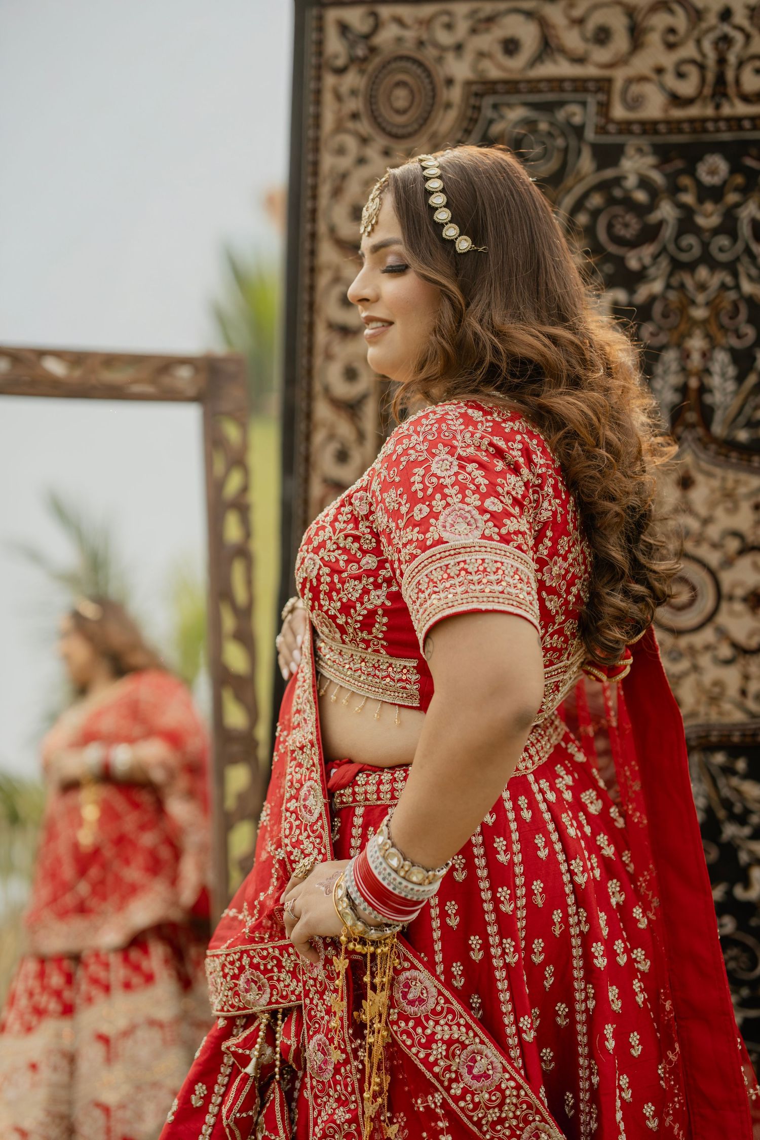 A bride in a red embroidered lehenga with gold jewelry and maang tikka poses elegantly near an ornate mirror.
