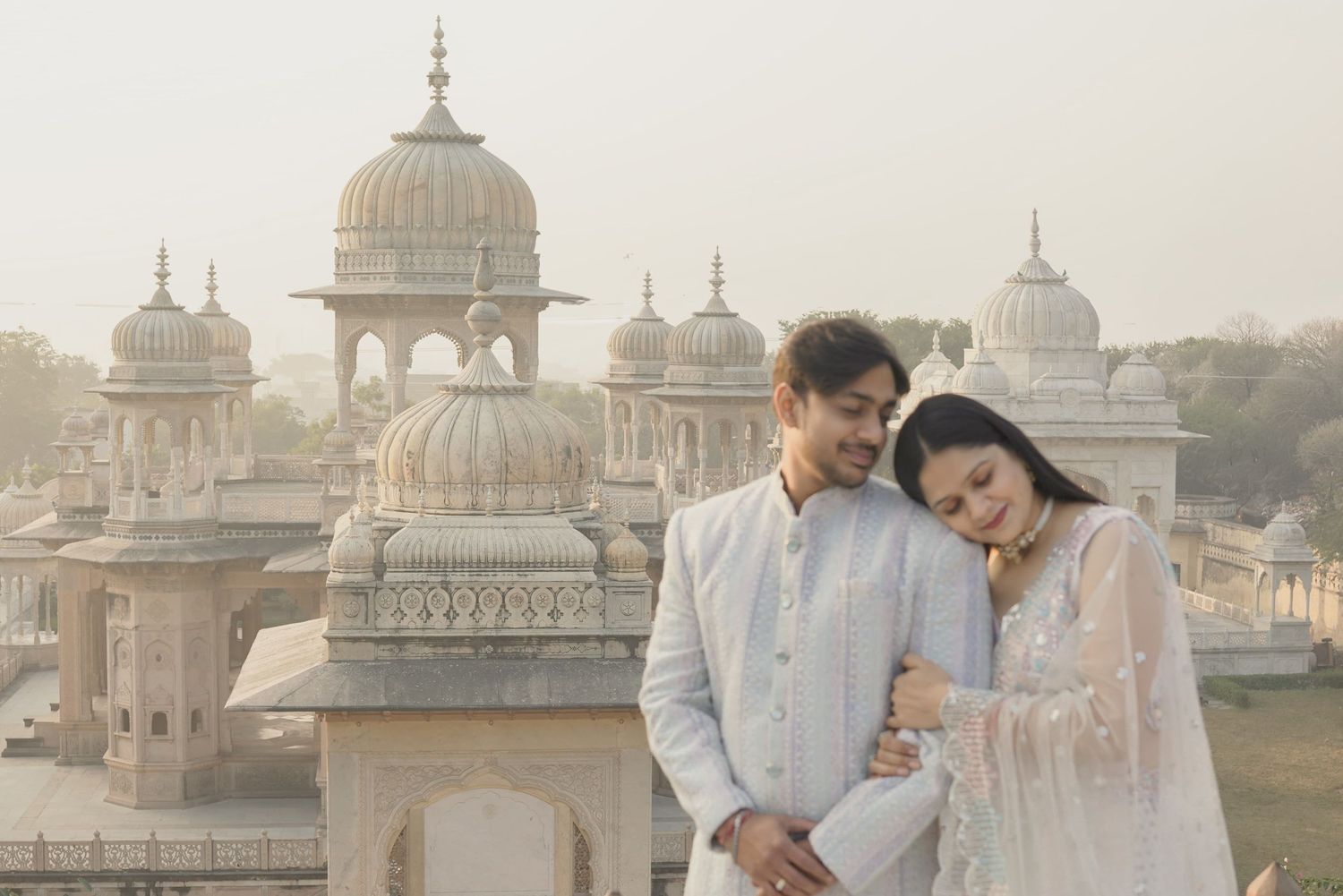 Couple in white traditional Indian attire posing at a historic Mughal monument with domed architecture.