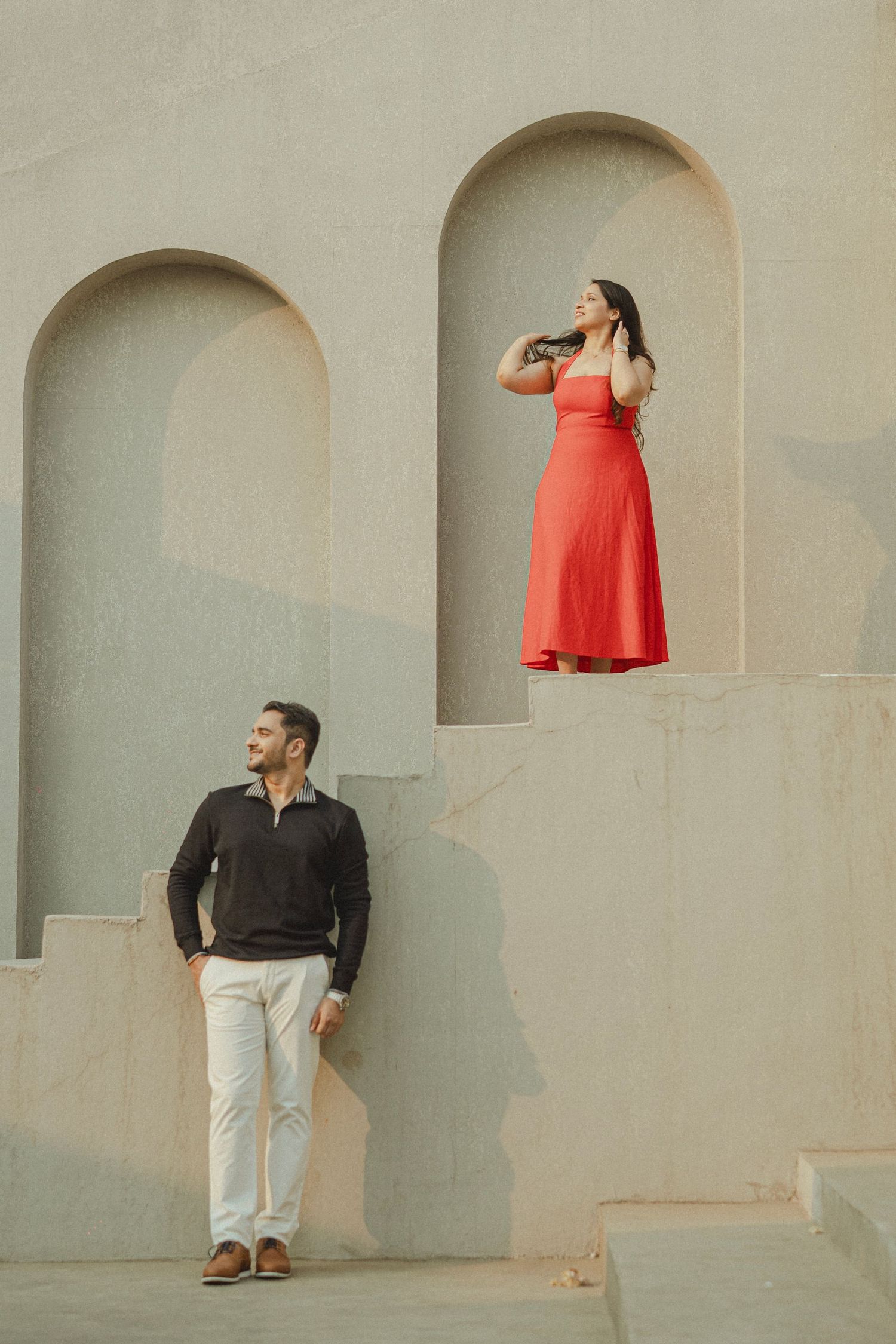 Man in black shirt and white pants looks up at woman in red dress standing in arched alcove of a beige wall.