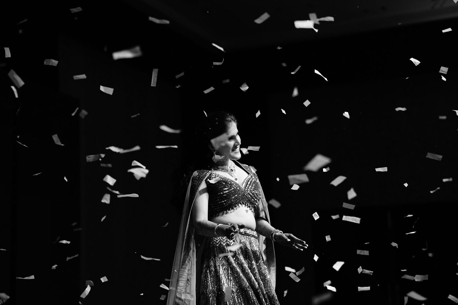 Black and white photo of a woman in a saree standing elegantly amid falling confetti at a celebration.