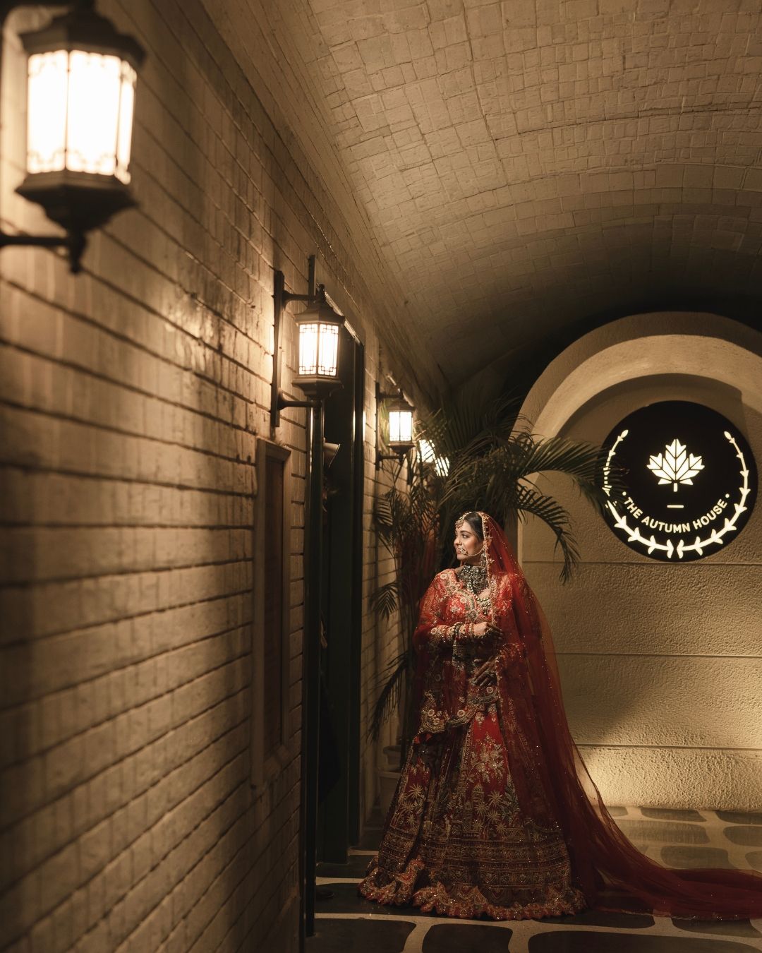 Bride in red bridal lehenga poses in arched corridor with vintage lanterns at The Autumn House venue.