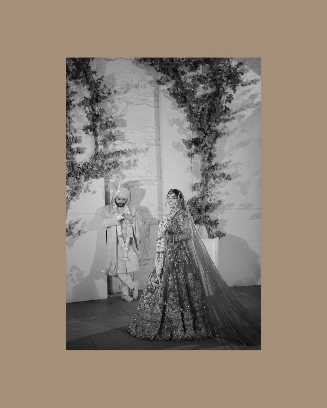Black and white photo of an Indian bride in ornate lehenga with veil and groom in sherwani posing outdoors.
