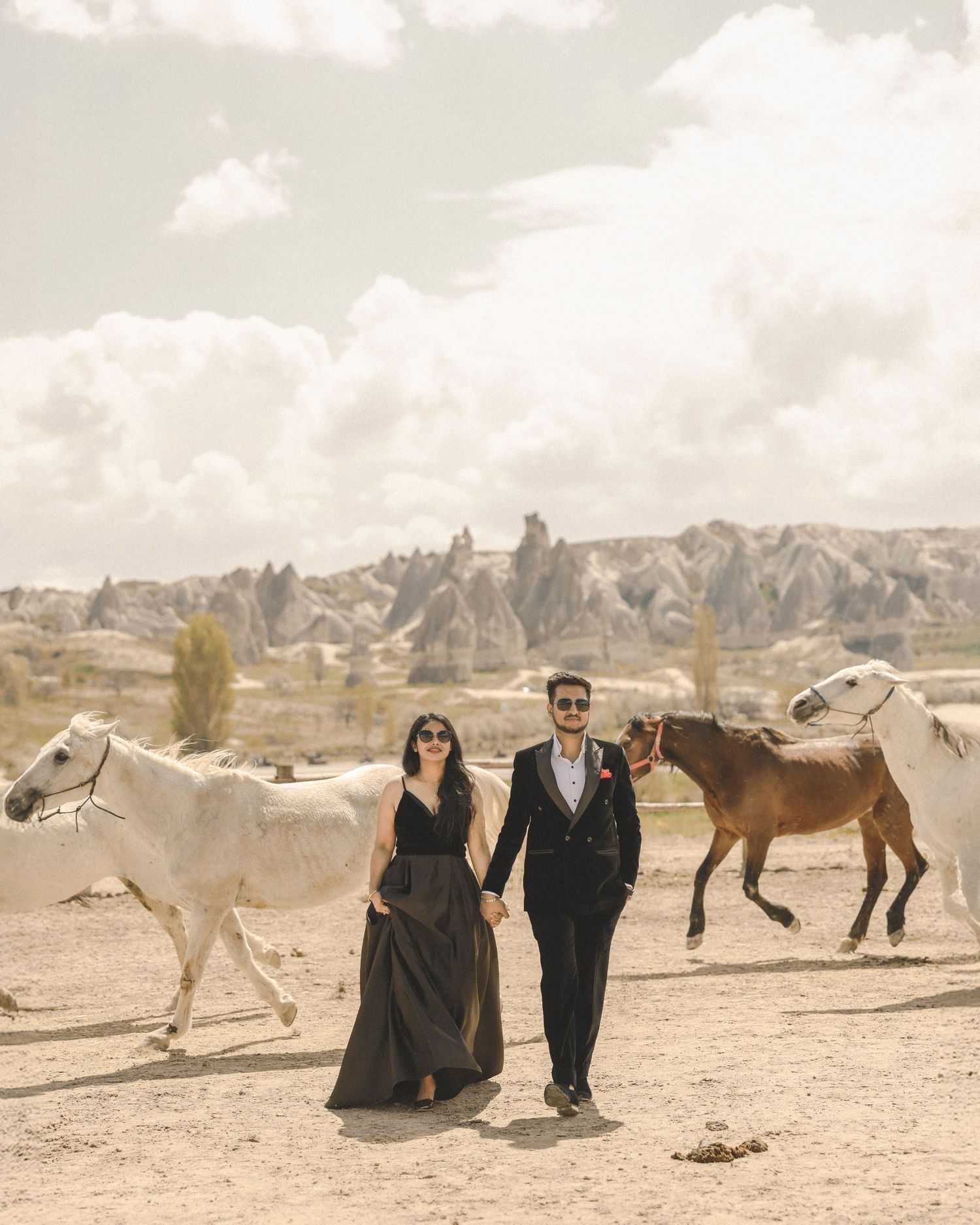 Couple in formal attire walks hand-in-hand among horses in Cappadocia, Turkey, with iconic fairy chimney rock formations behind.