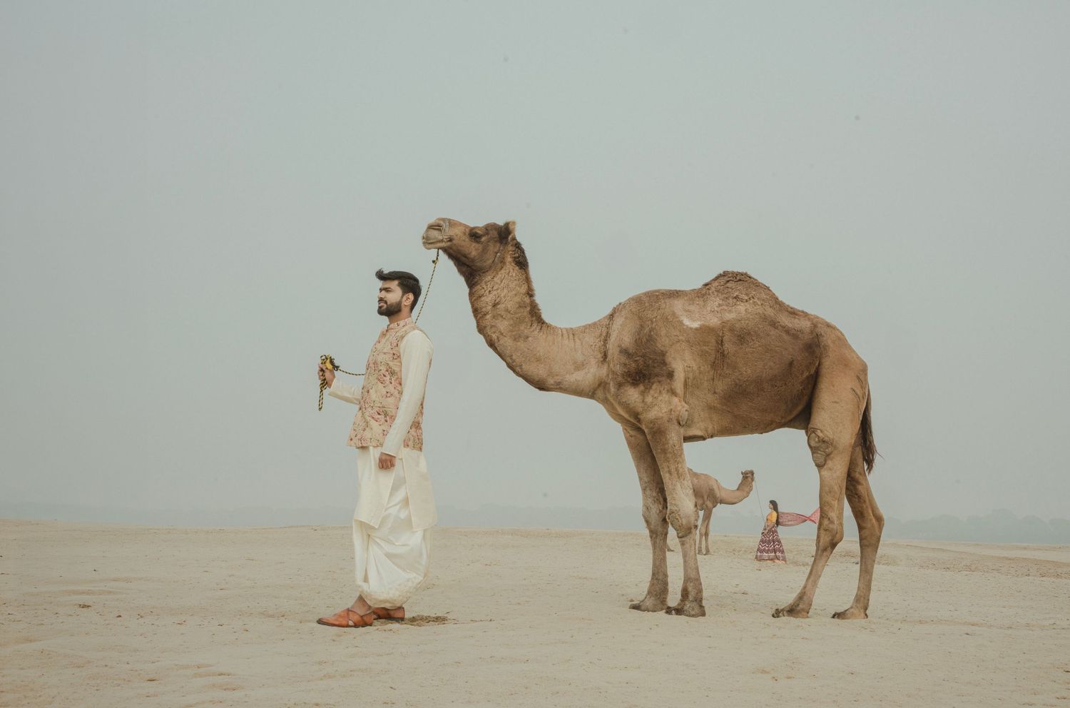 A man in traditional white robes stands beside a camel on a sandy desert landscape under a hazy sky.