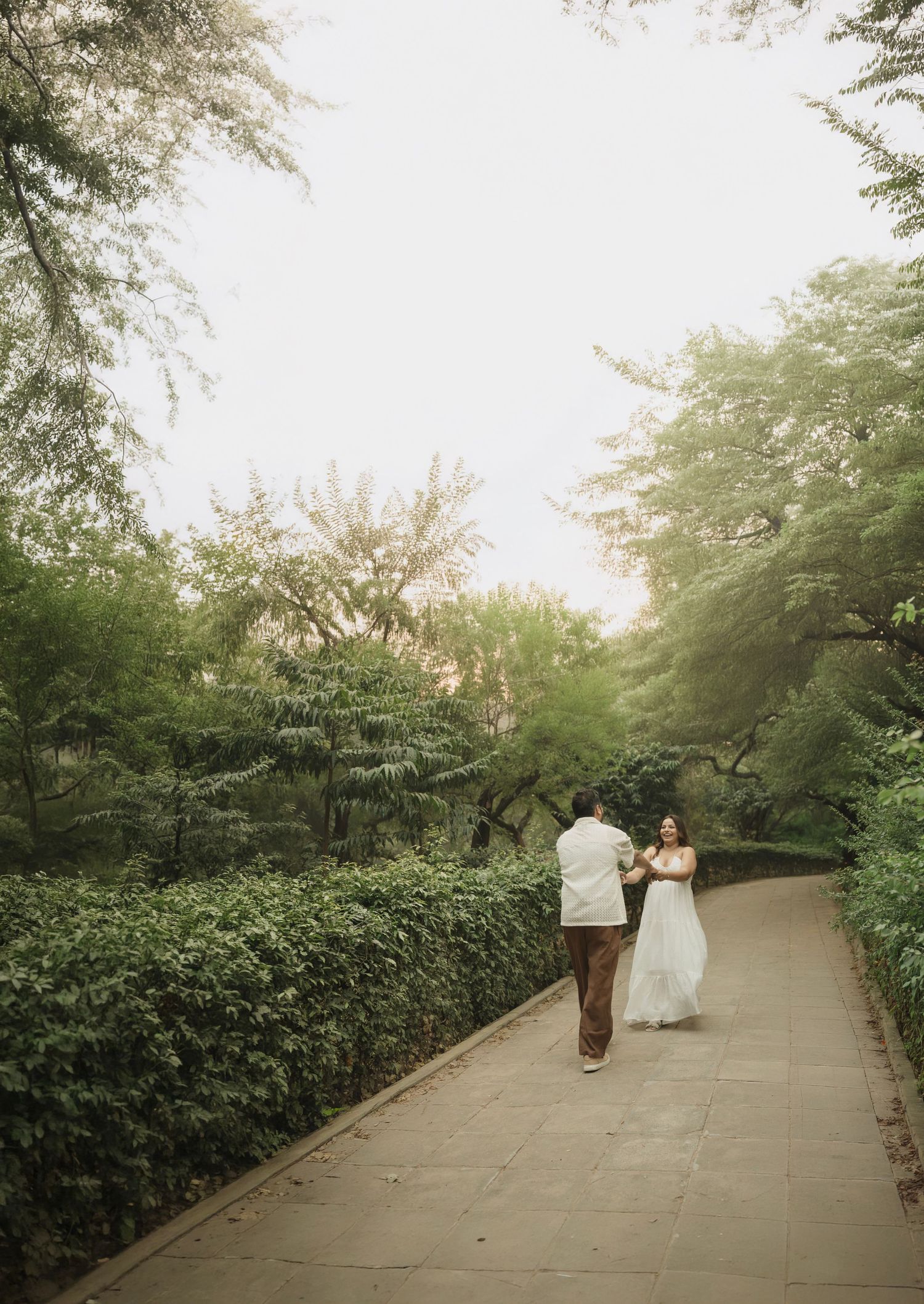A bride and groom walk hand in hand along a lush green tree-lined garden path on their wedding day.