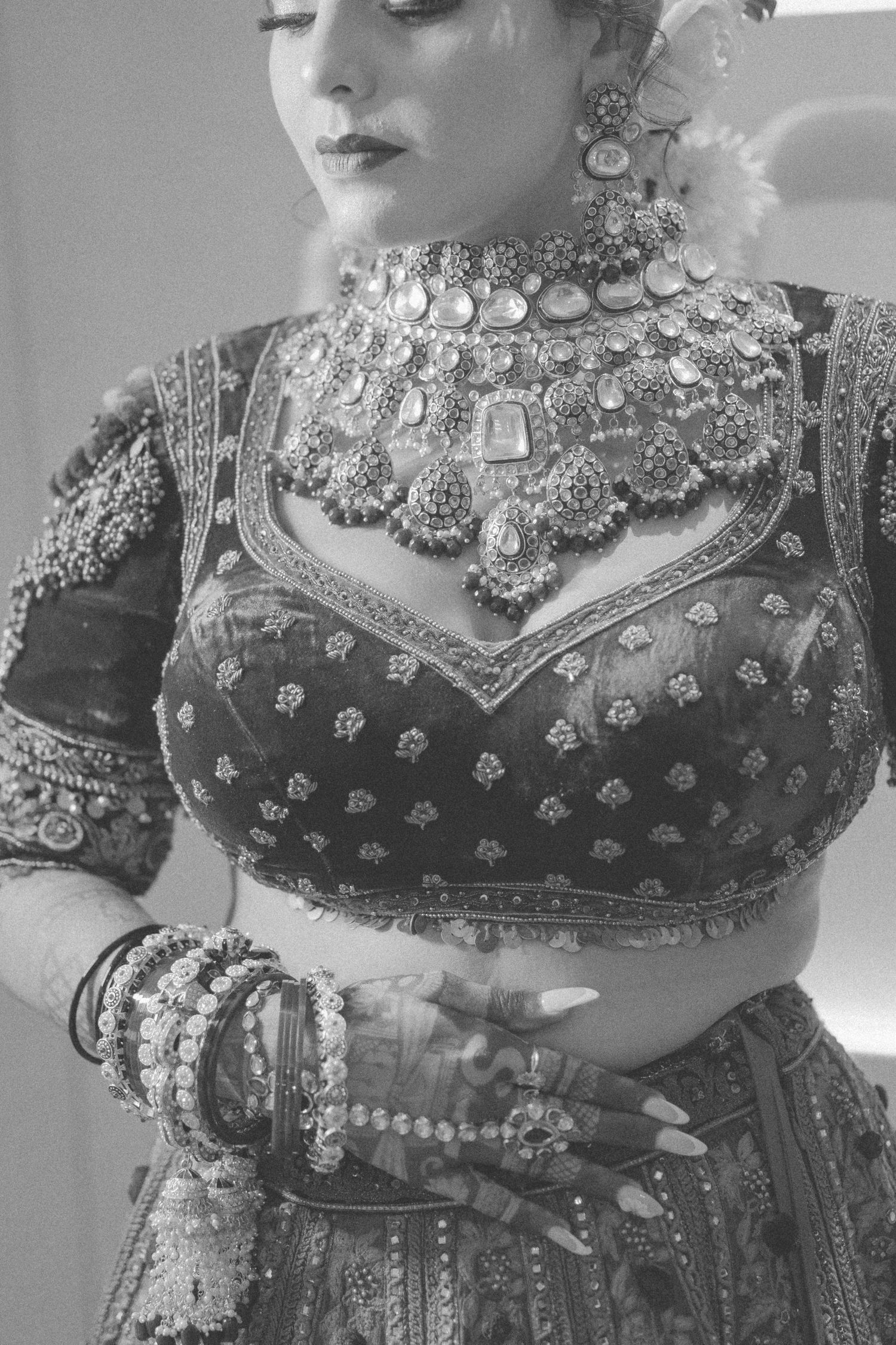 Black and white photo of a bride in embroidered lehenga choli with ornate necklace, bangles, and henna on hands.