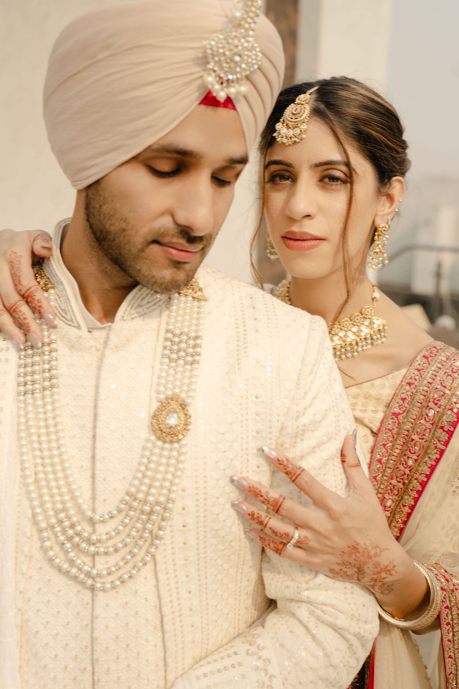 Sikh bride and groom in traditional ivory wedding attire with gold jewelry and pearl necklace posing together.