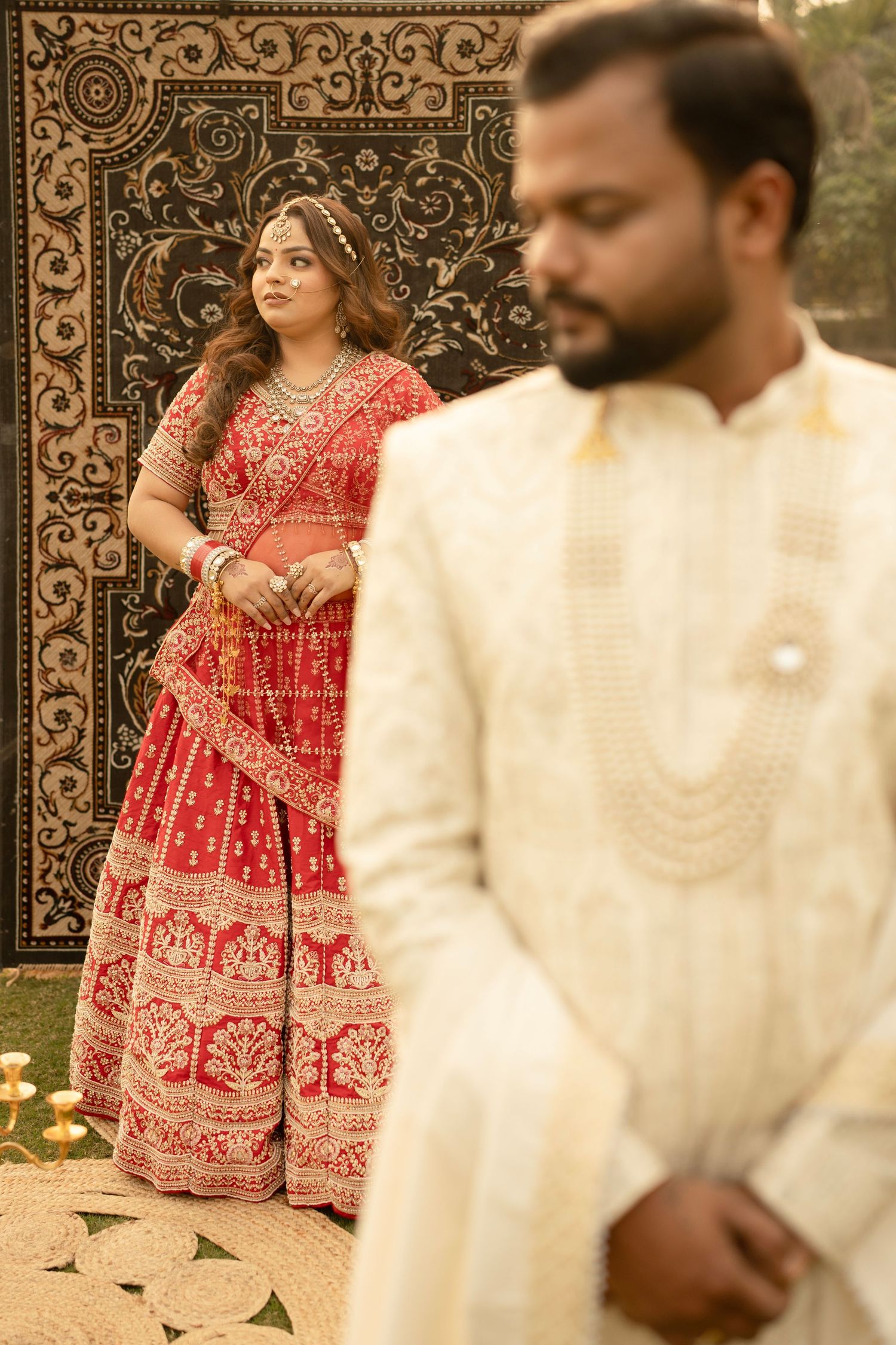 Indian bride in red lehenga stands before ornate backdrop as groom in cream sherwani poses in foreground.