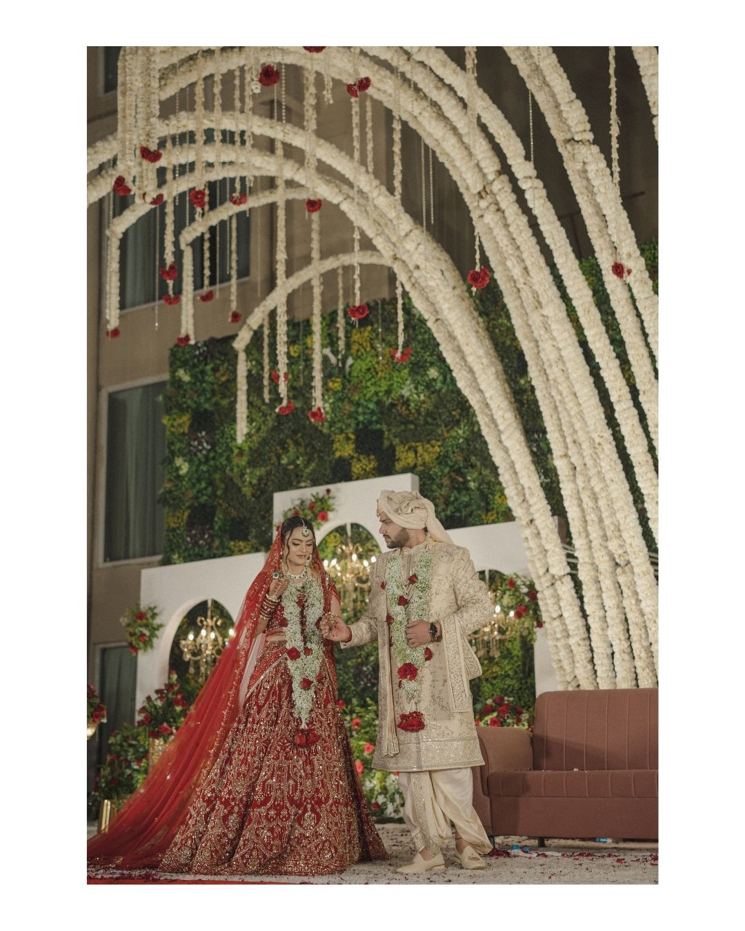 Indian bride in red lehenga and groom in ivory sherwani pose under floral arches adorned with white and red flowers.