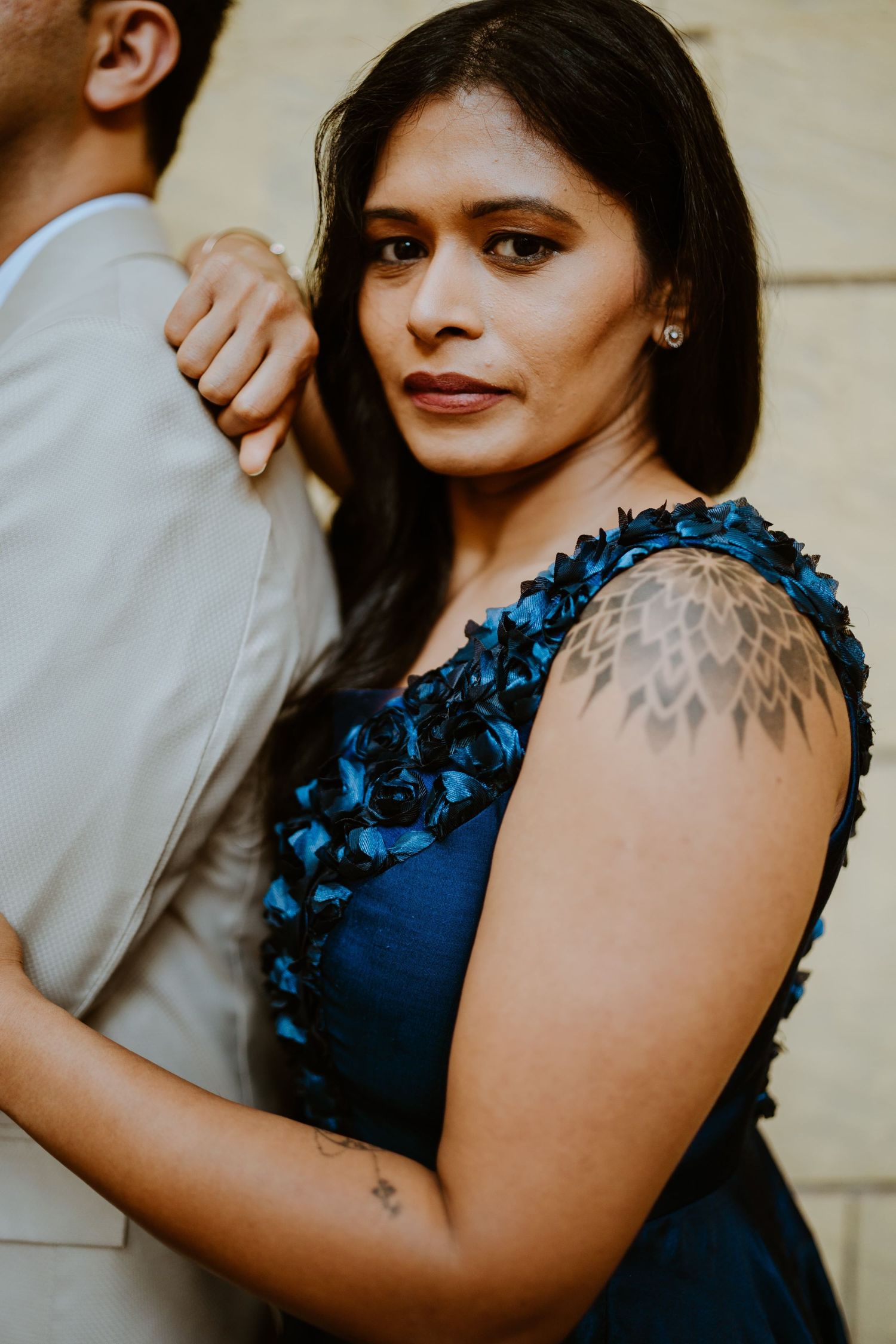 Woman in blue floral dress with shoulder tattoo embraces man in suit, gazing at camera in elegant portrait.