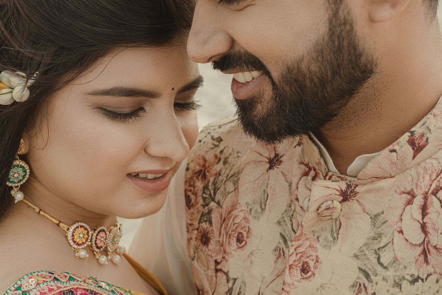 A close-up of an Indian couple in traditional attire sharing a romantic moment, smiling and dressed in floral wedding wear.