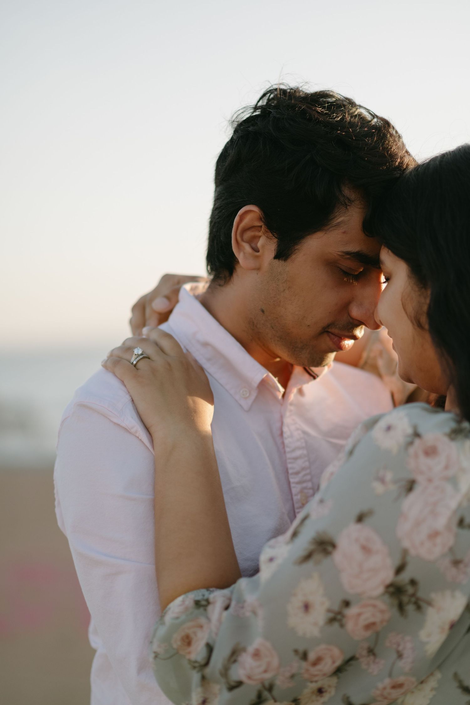 A couple shares a tender romantic moment at the beach, with the woman wearing a floral dress and engagement ring.