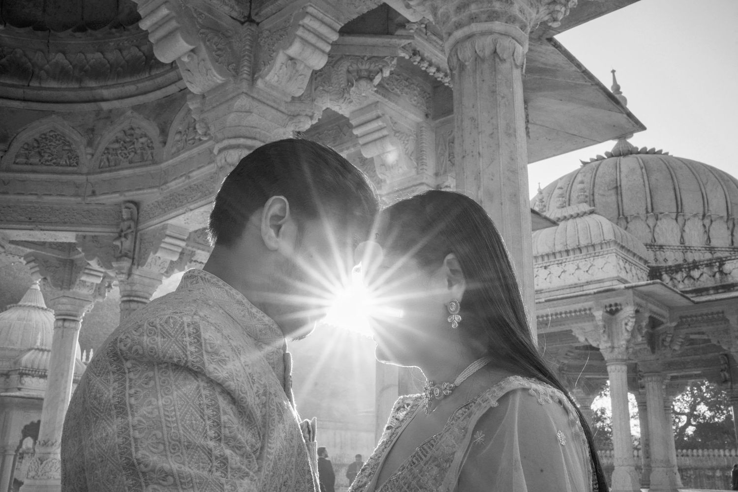 Black and white photo of a couple facing each other with a sunburst between them at an ornate Indian temple.