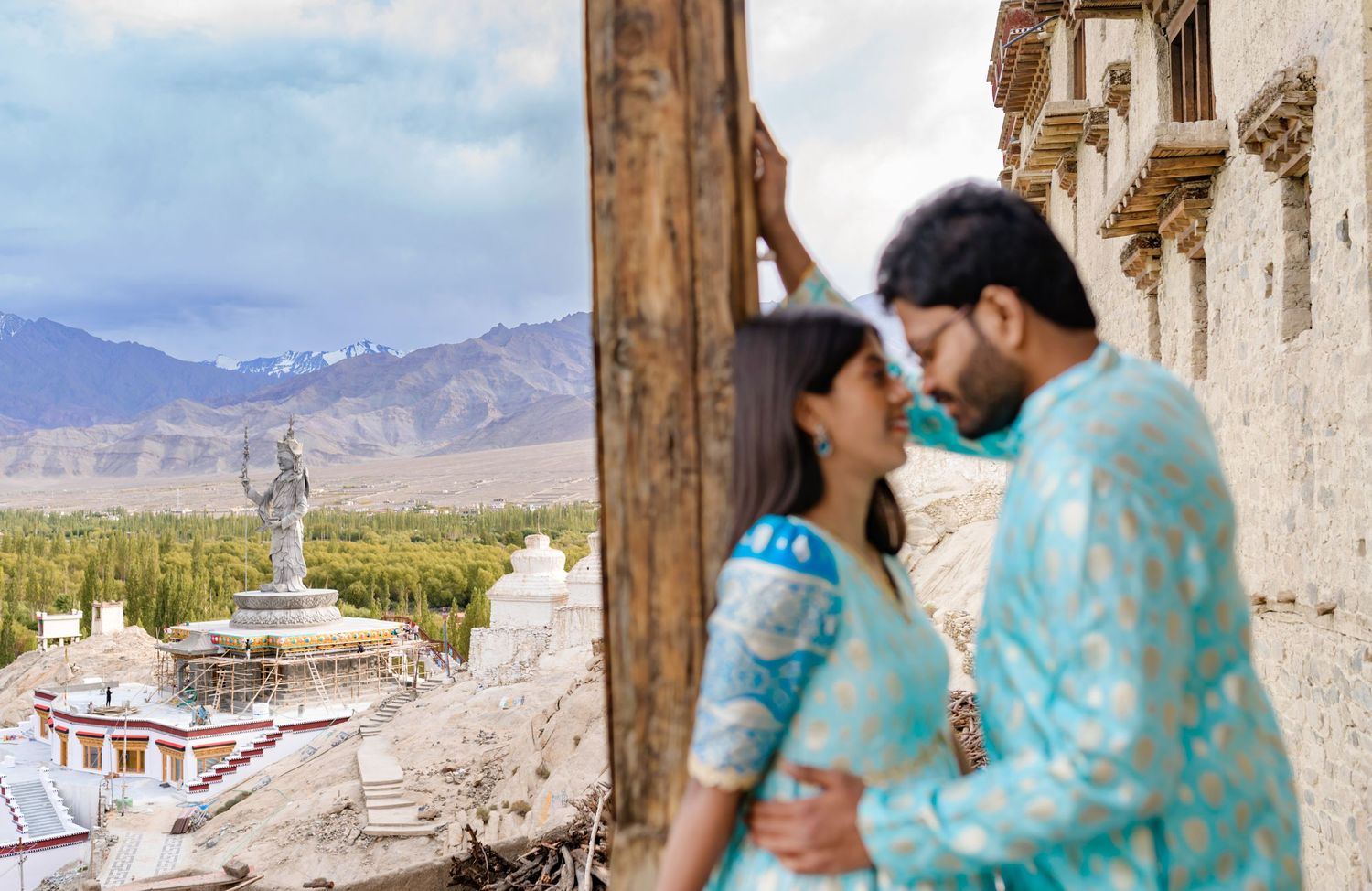 Couple in matching turquoise outfits at Leh Ladakh monastery with mountain valley and Buddha statue backdrop.
