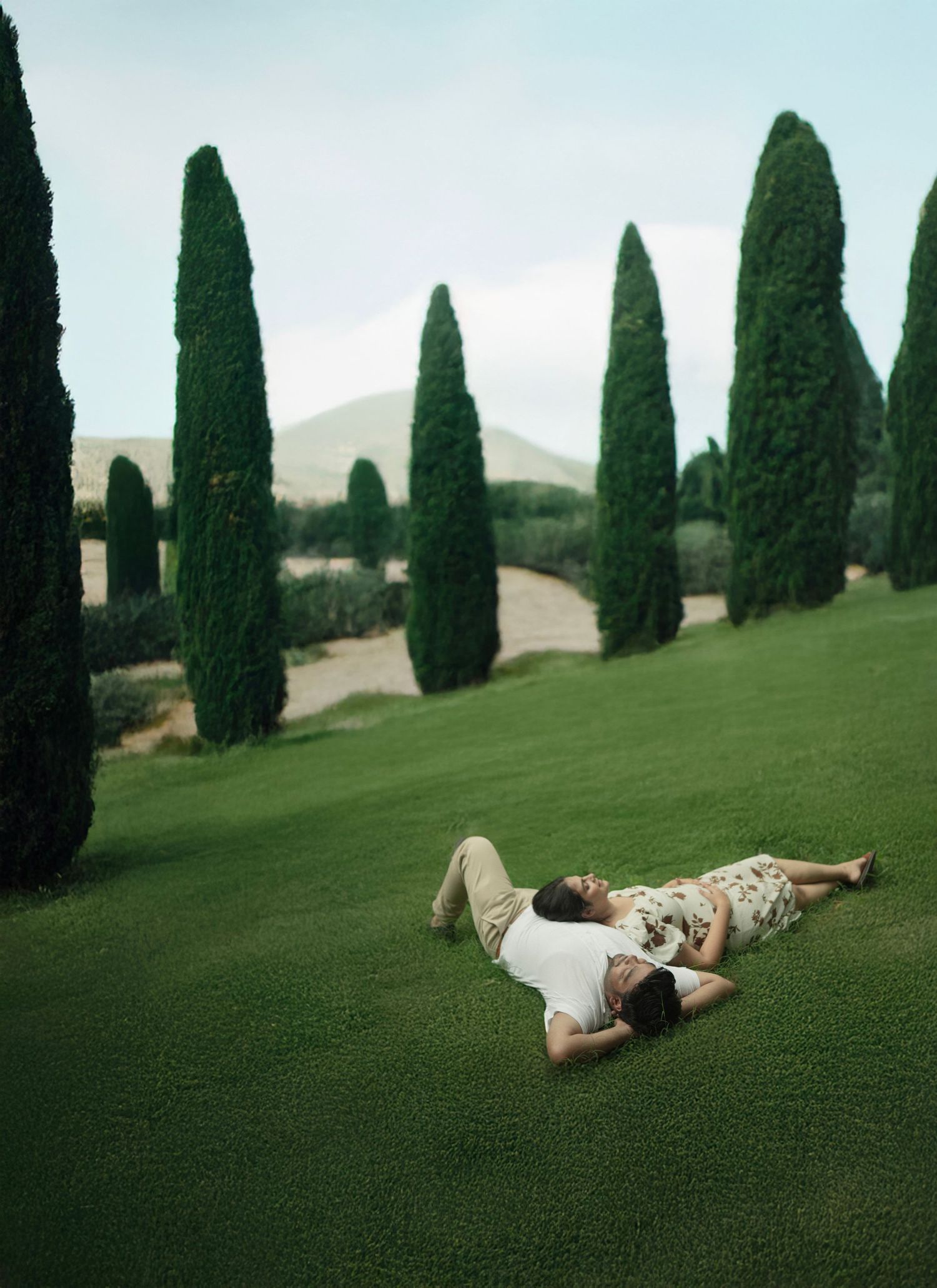A couple lying together on lush green grass surrounded by tall cypress trees in a scenic landscape.
