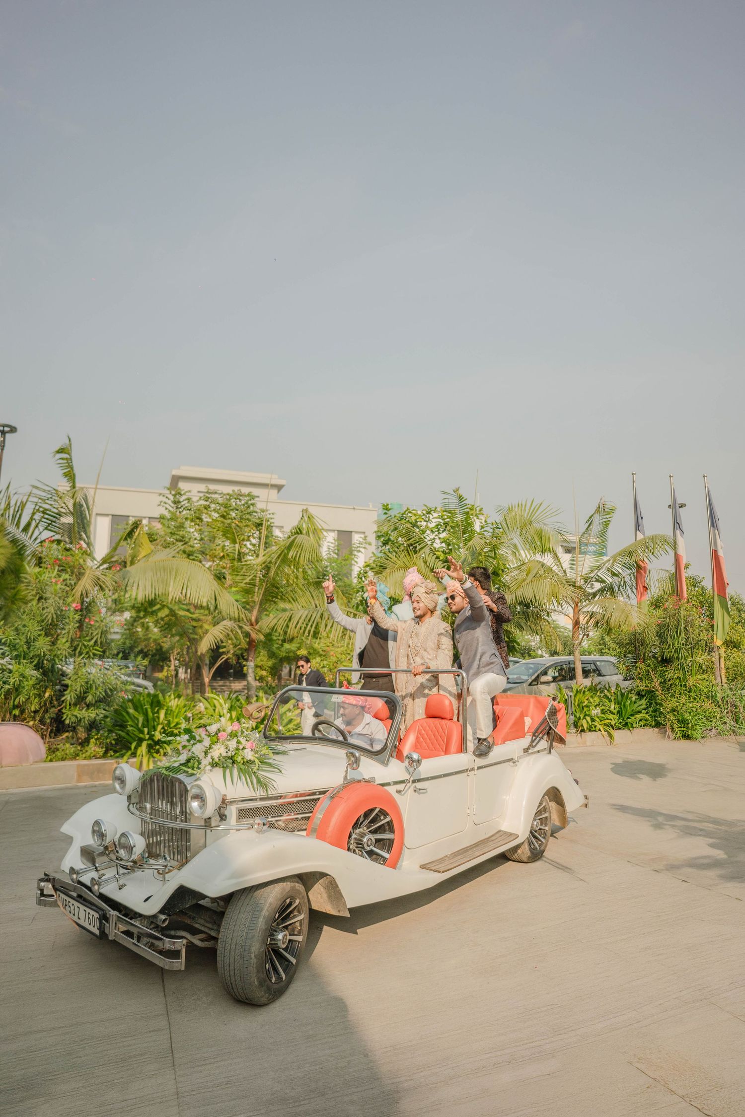 Two people standing in a vintage white convertible car with red interior, celebrating at an outdoor wedding venue.