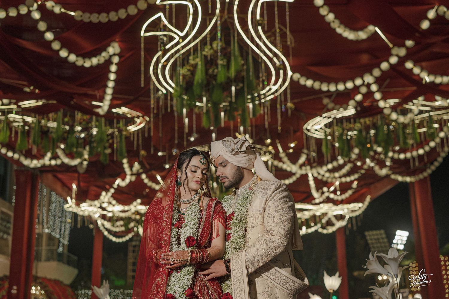Indian bride and groom in traditional wedding attire under floral decorations and fairy lights at an elegant ceremony.