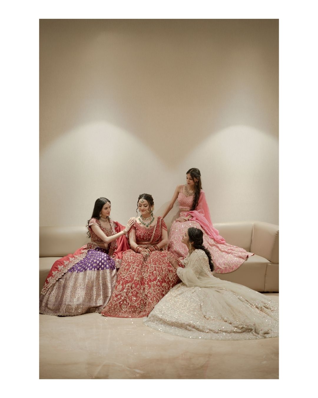 Four women in vibrant Indian bridal lehengas pose together in an elegant beige studio setting.
