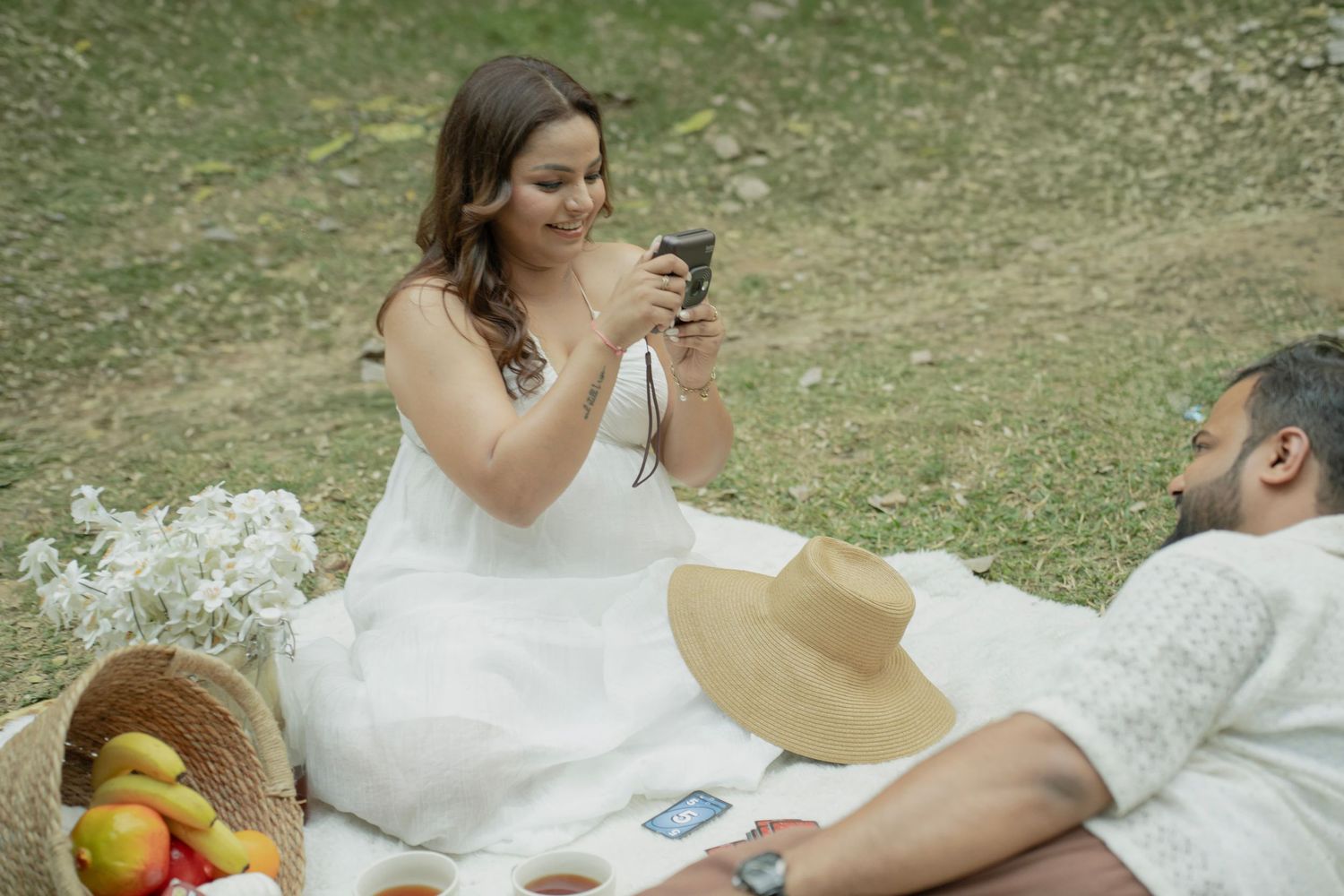 Couple enjoying a romantic outdoor picnic, woman photographing man lying on blanket with fruits and hat nearby.