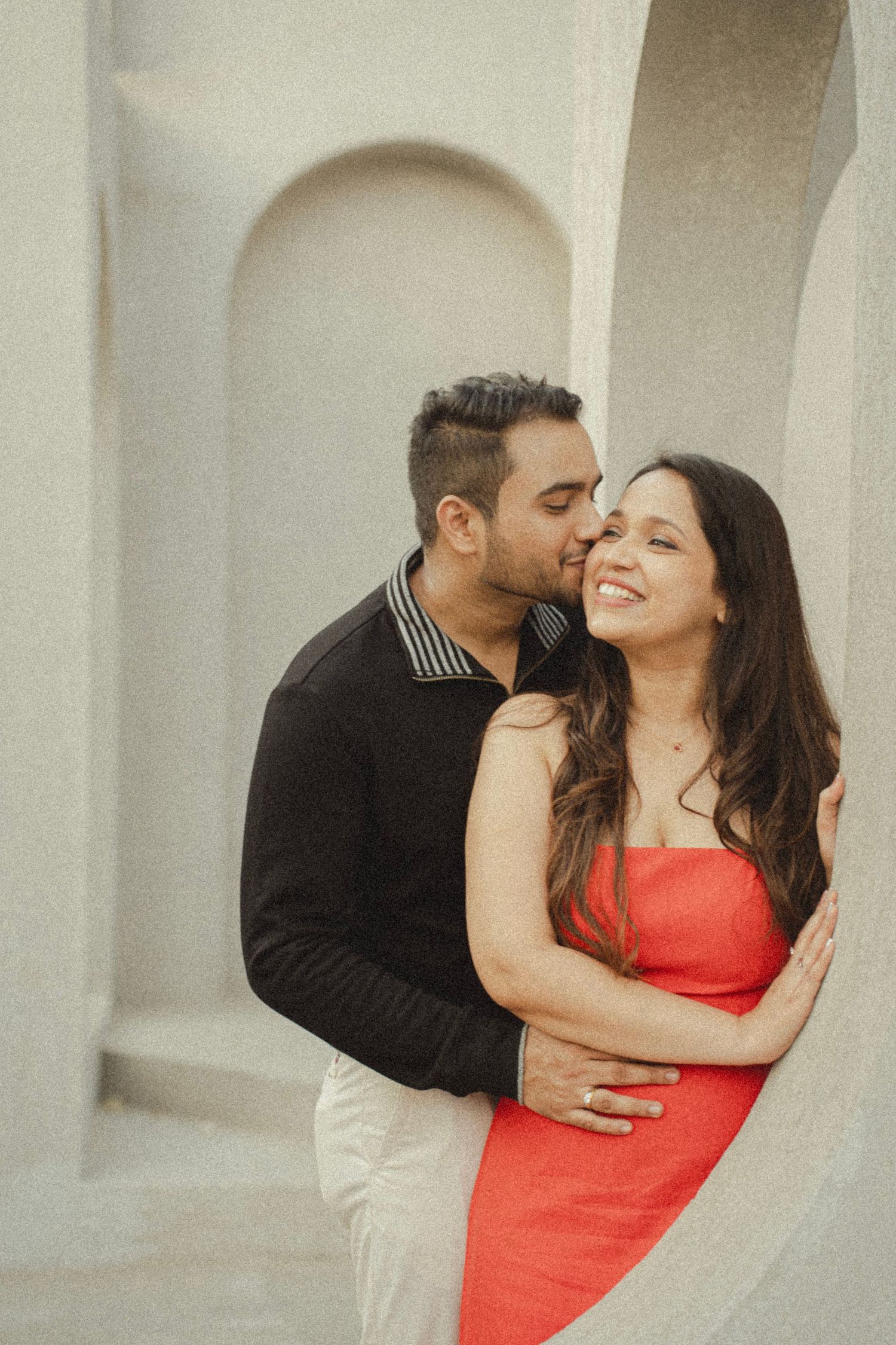 A couple shares a romantic moment, man in black shirt kissing woman in red dress against white arched wall.