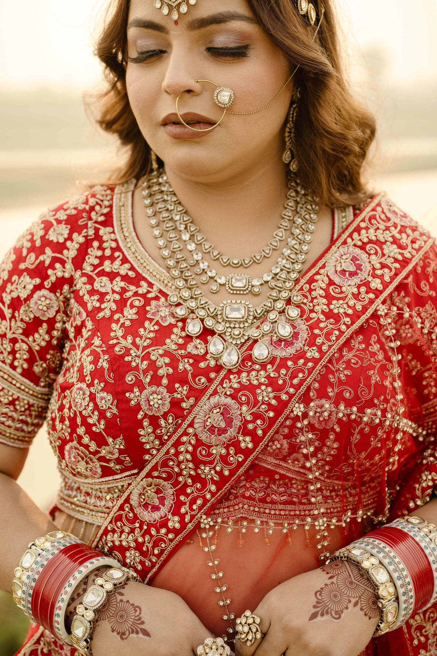Close up of traditional Indian bridal jewelry and red embroidered lehenga with intricate gold details and mehndi adorned hands.