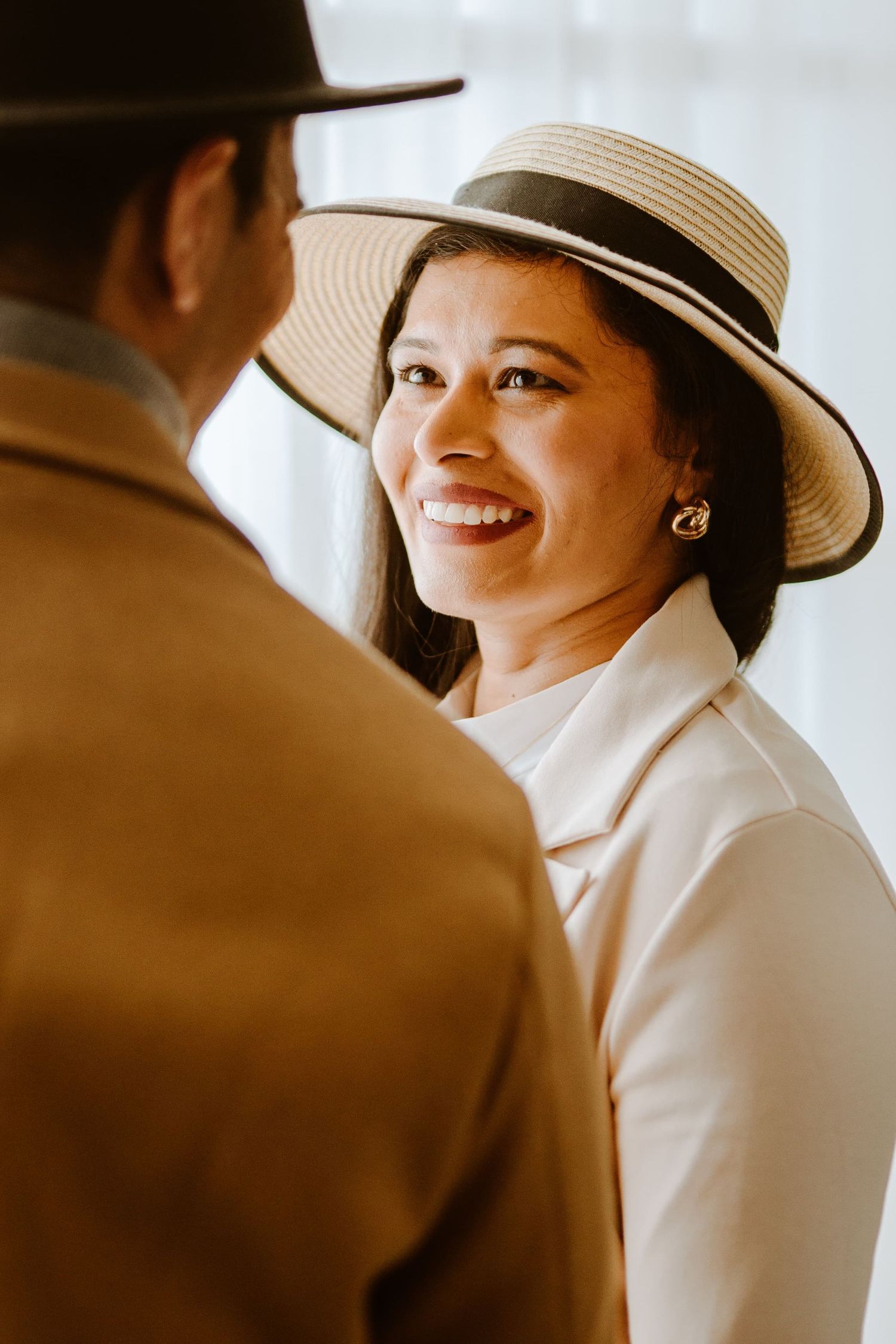 A smiling woman wearing a straw boater hat and white jacket looks up at a man in a brown coat and hat.