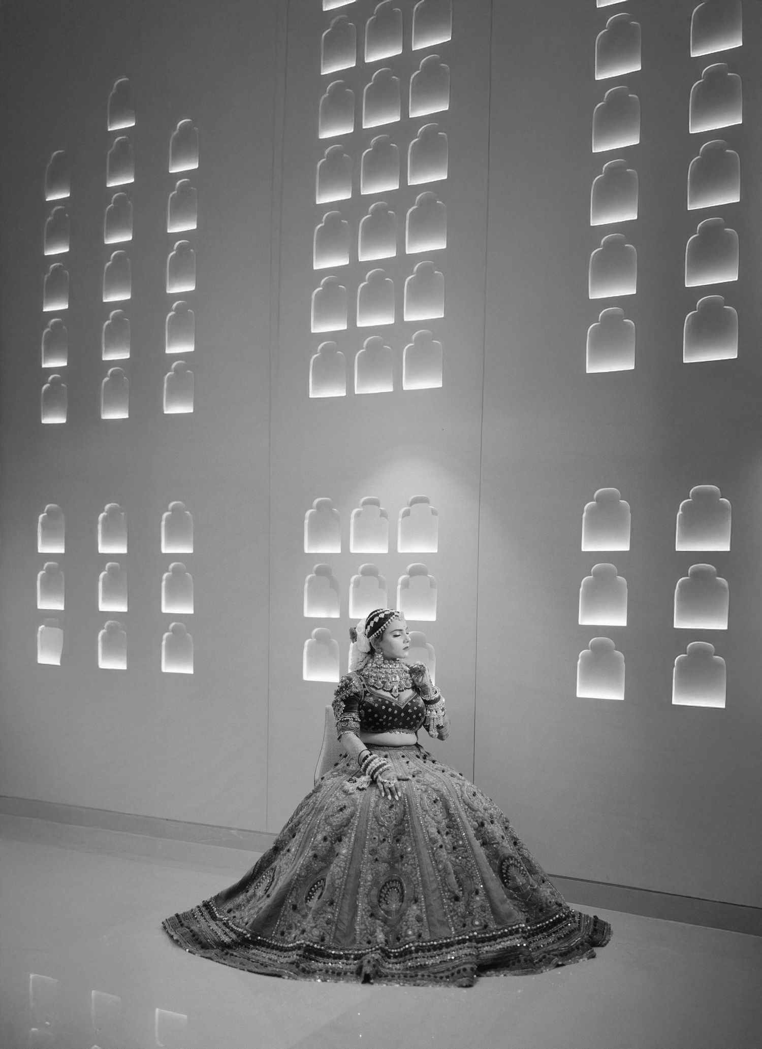 Black and white photo of a bride in an elaborate lehenga seated before an ornate illuminated wall with arched niches.