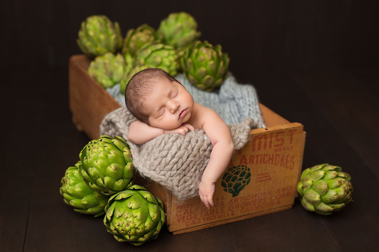 Sleeping newborn nestled in wooden crate surrounded by fresh green artichokes on dark background.