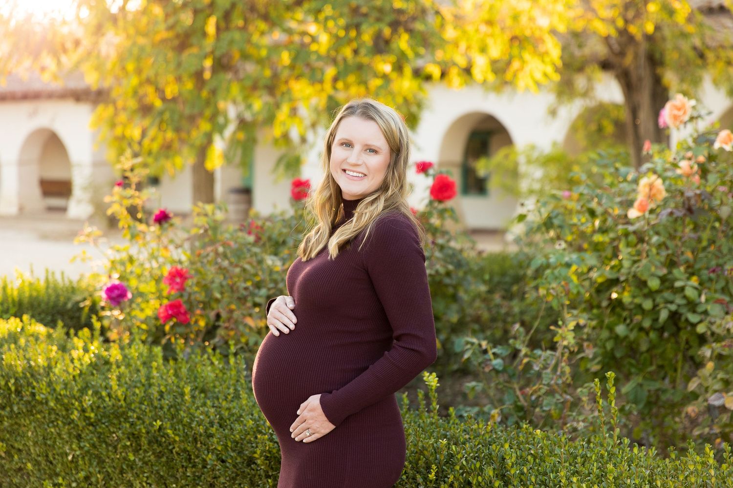 Maternity portrait in garden with burgundy dress against blooming roses and autumn foliage.