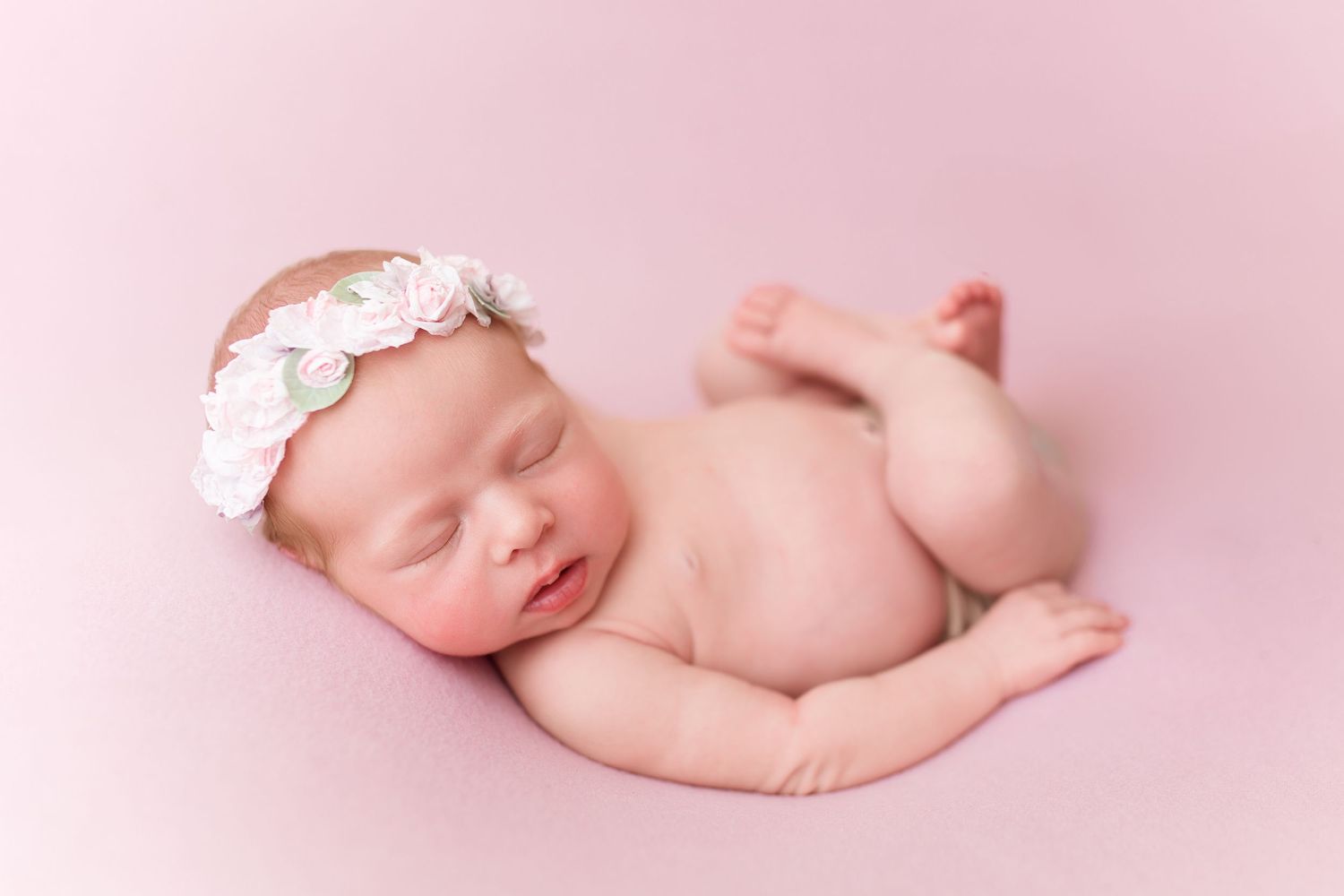 Sleeping newborn wearing floral headband posed on pink background.