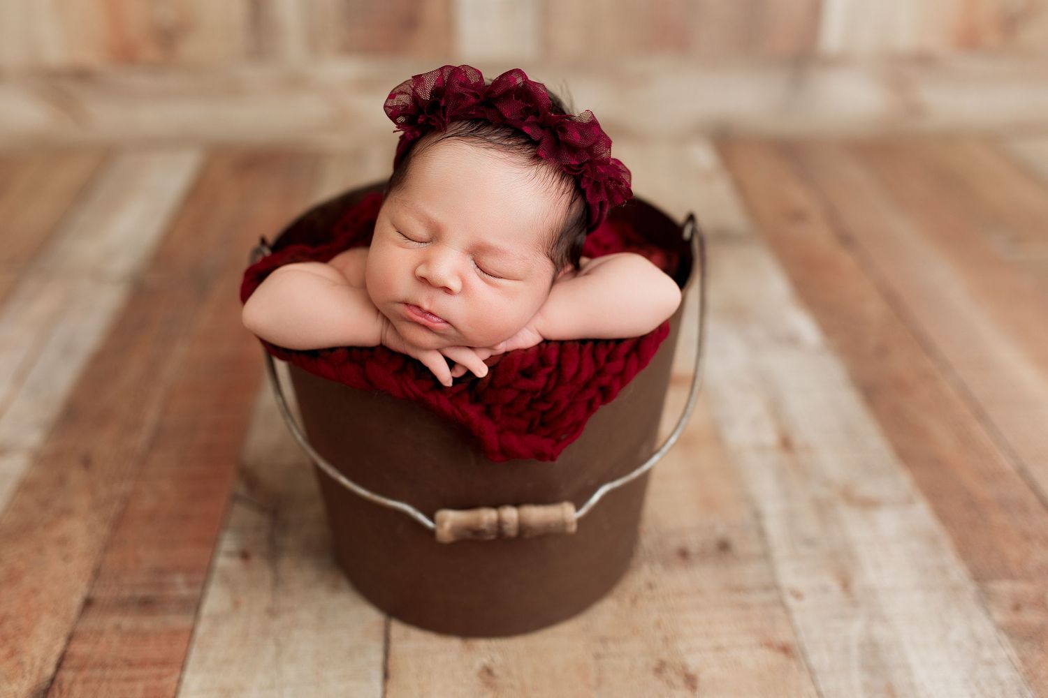 Sleeping newborn baby posed in rustic brown bowl with red knit blanket.