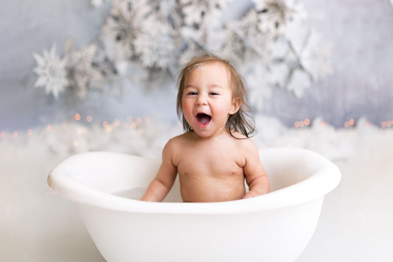 Baby laughing in white bathtub with winter snowflake backdrop.