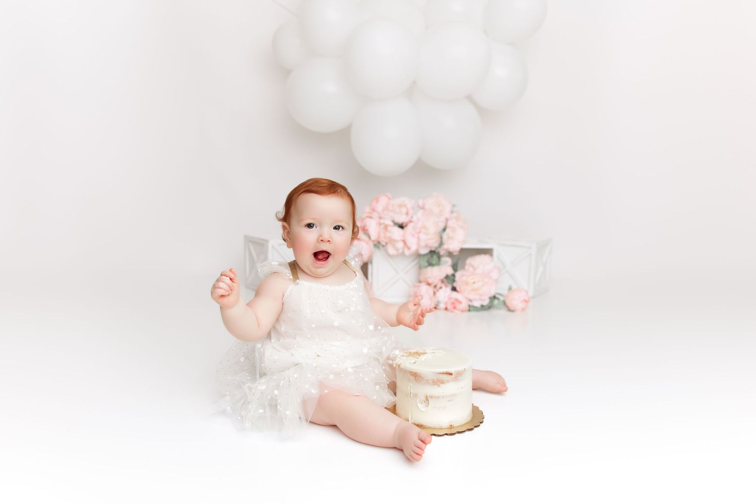 White studio setup with baby in white outfit surrounded by balloons and soft toys.