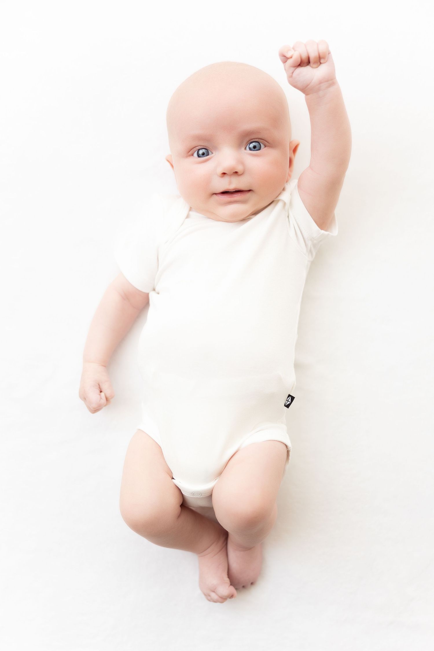 Baby in white bodysuit poses against pure white backdrop in minimalist style.