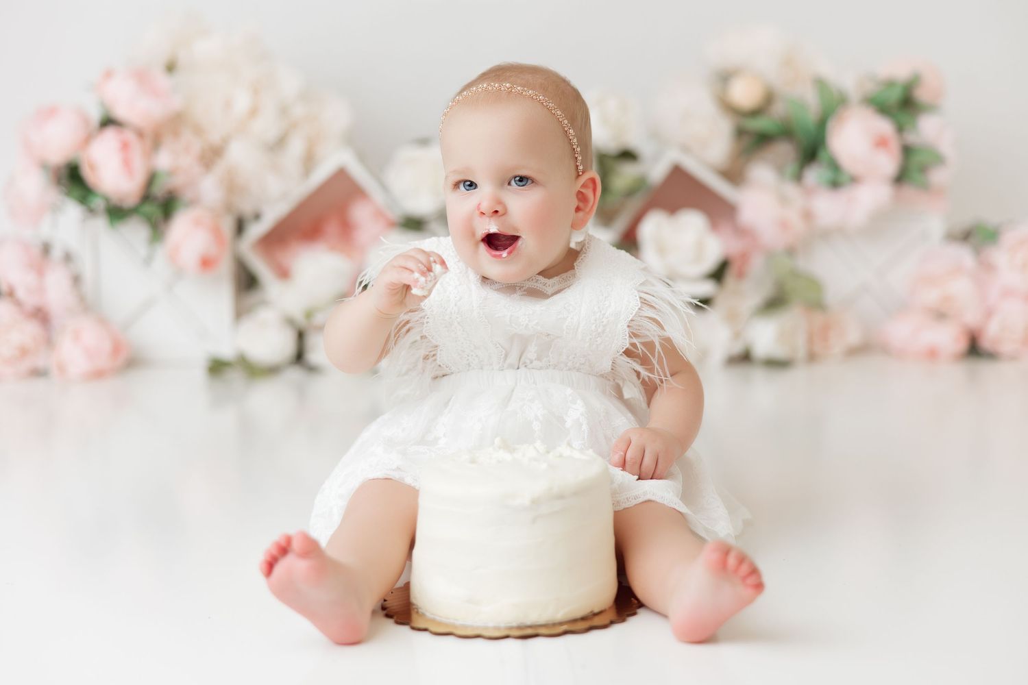 Baby in white dress celebrating first birthday with cake and pink floral decorations.