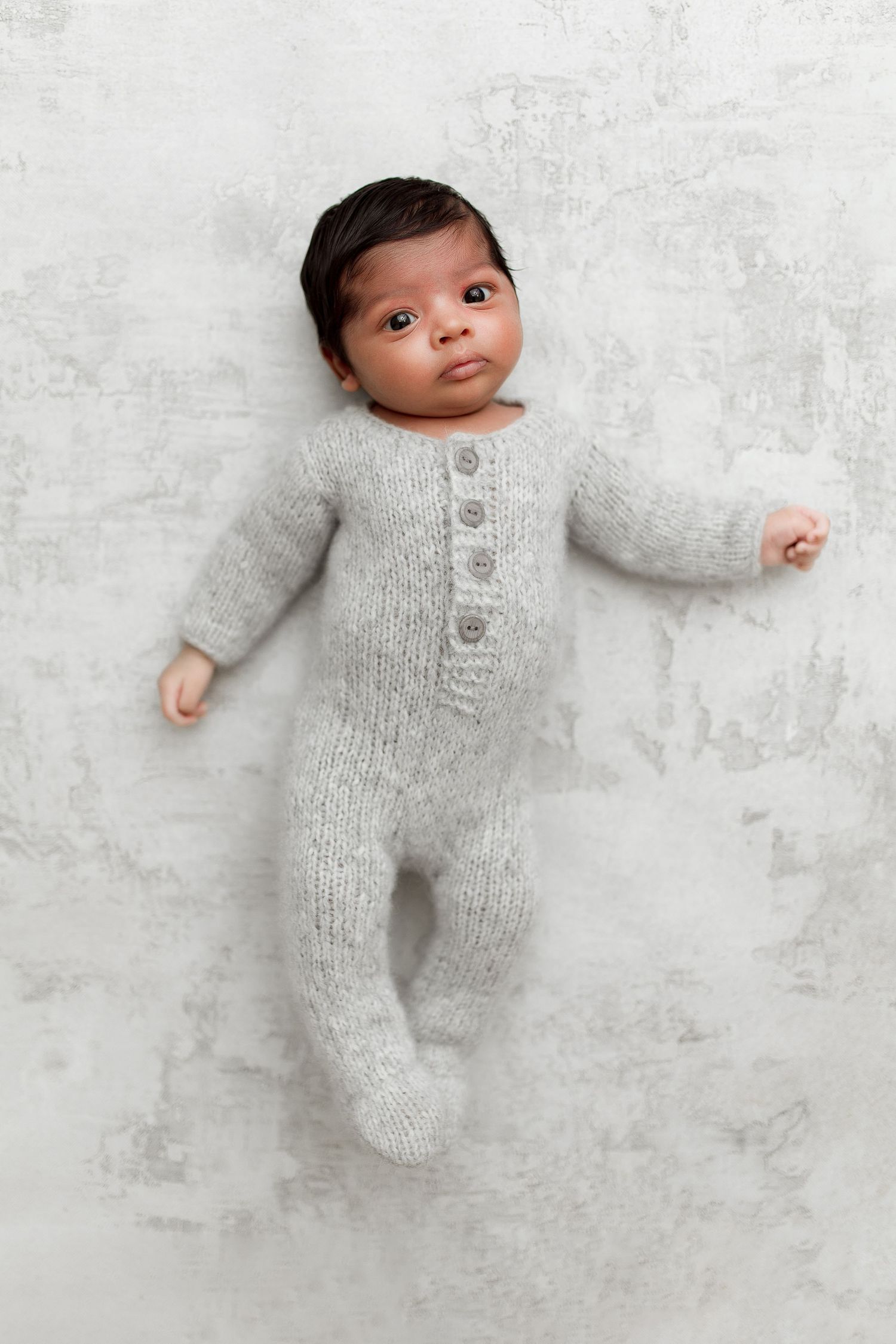 Newborn wearing cozy gray knit romper lying on white textured background in artistic photo series.