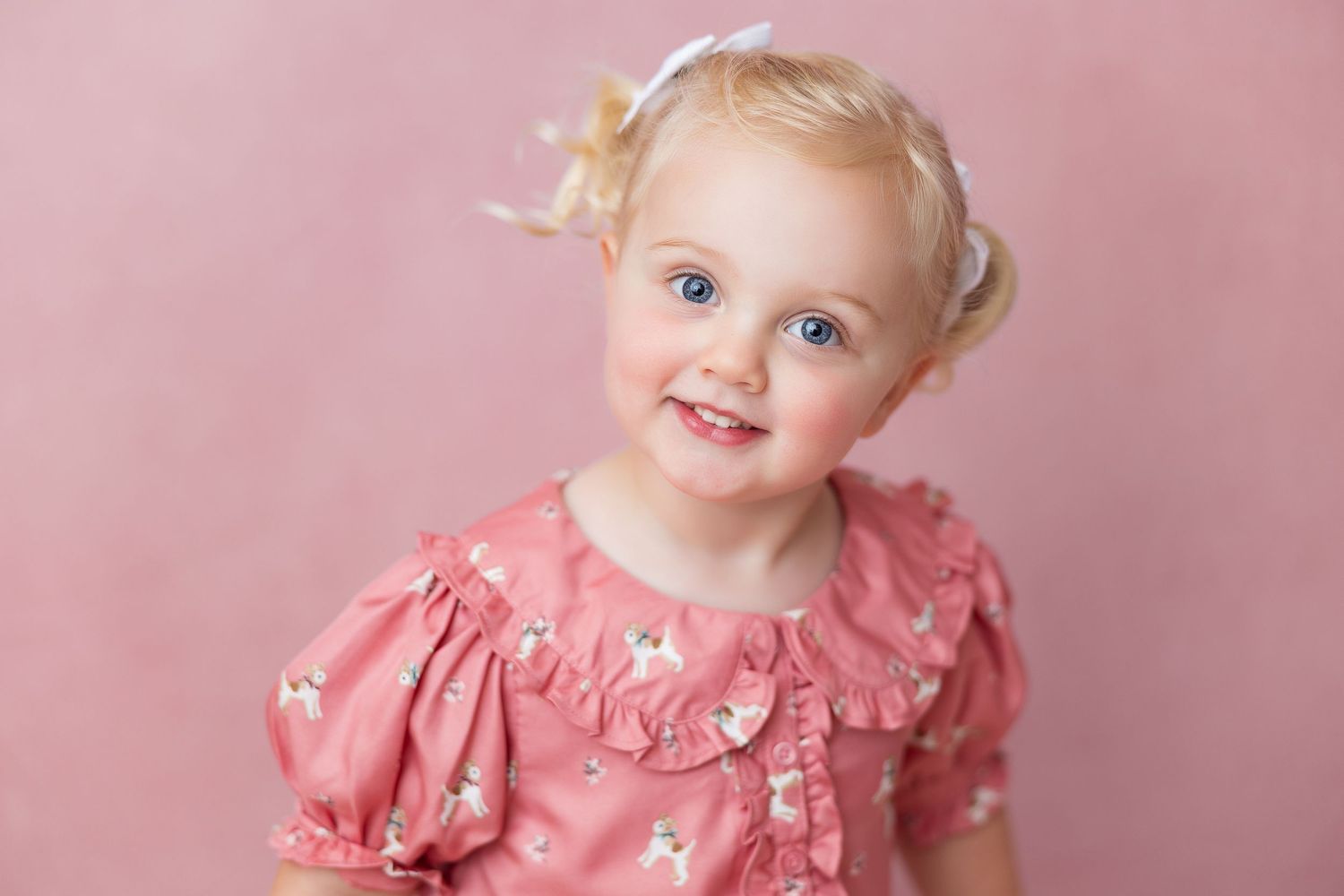A young child in a pink dress with ruffled sleeves sits against a soft pink studio backdrop.