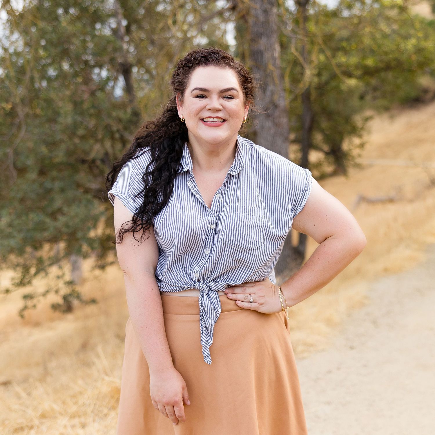 Someone wearing a striped tie-front top and peach-colored skirt poses outdoors on a dirt path in a natural setting.