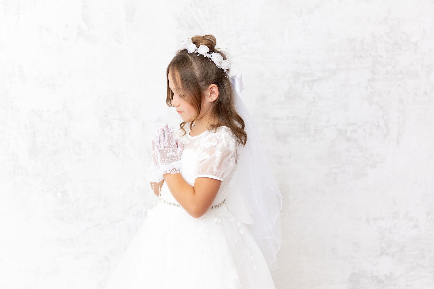 Young girl in a white lace dress and floral hairpiece stands against a textured white wall.