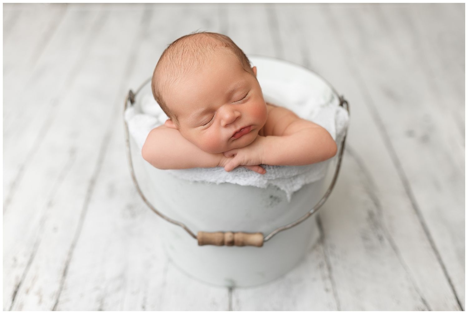 Sleeping newborn baby wrapped in white blanket rests peacefully on light wooden floor during professional photoshoot.