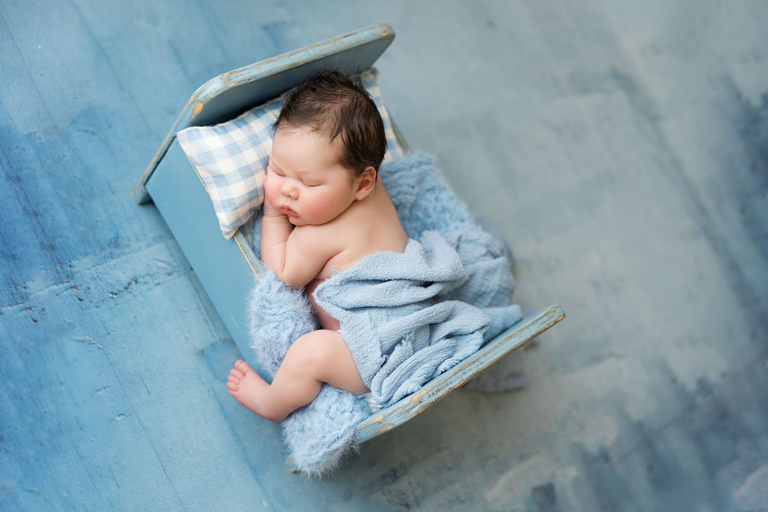 A sleeping baby in light blue swaddle blanket lies on a textured blue backdrop during a newborn photo session.