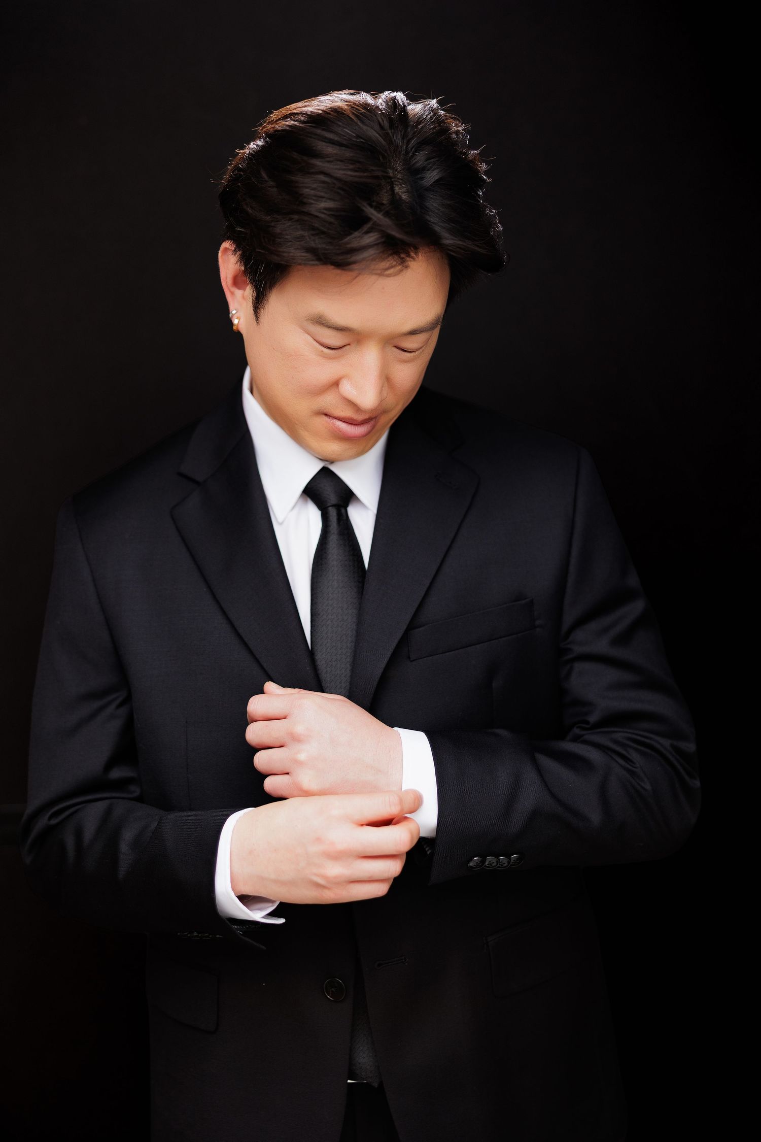 Asian man in black suit adjusting cufflinks against dark background, professional business attire.