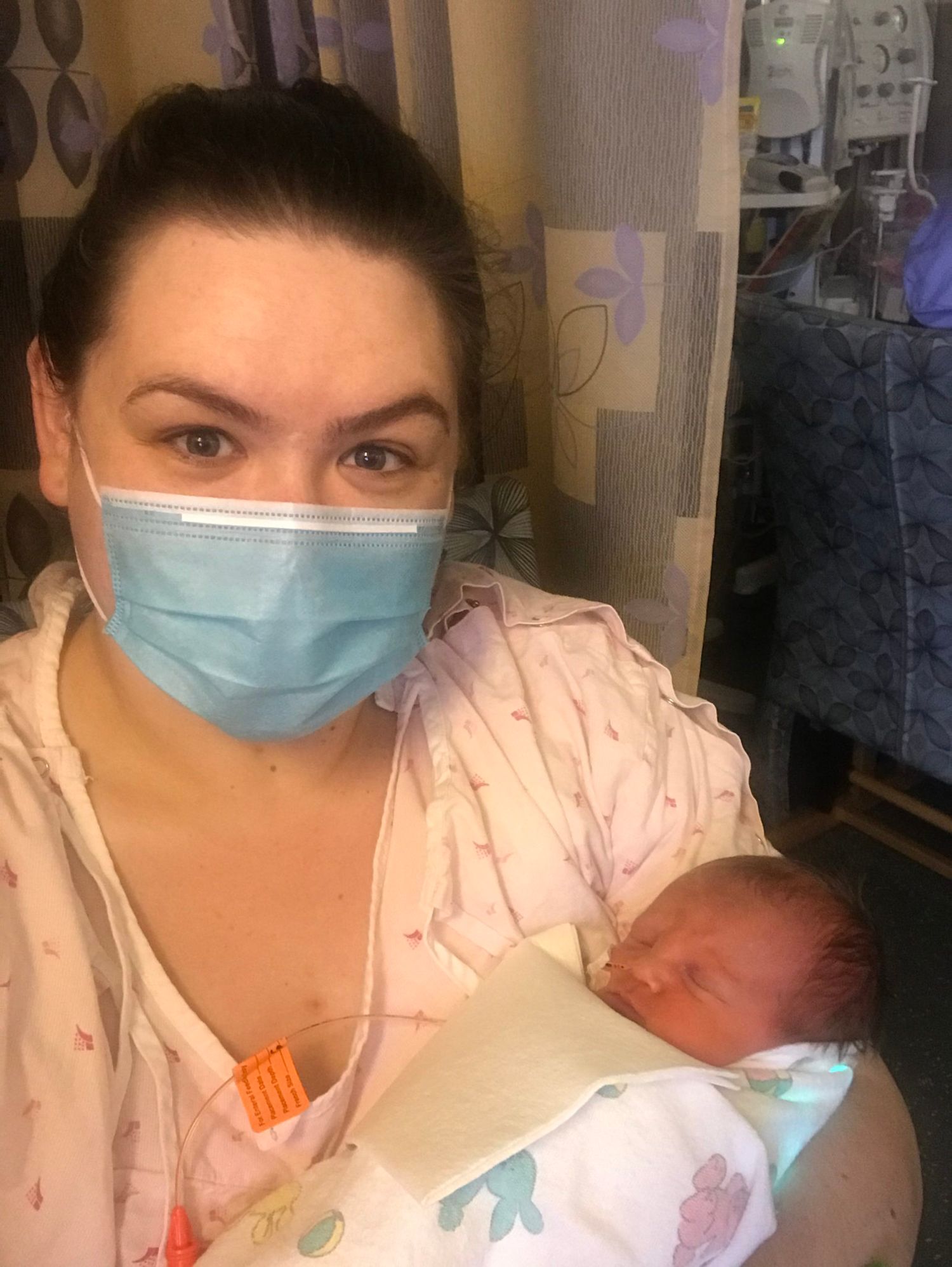 Healthcare worker in protective mask holds a newborn baby while sitting in a medical setting.